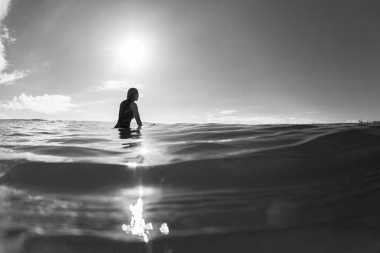 Girl Surfer Silhouetted Sitting on surfboard waiting ocean water in black and white lifestyle photo.