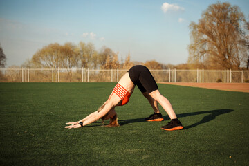 Sporty woman is practicing yoga, standing in downward-facing dog outdoors on sports ground in spring