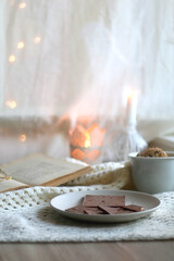 Fototapeta na wymiar Plate with almond chocolate bar, mug with chocolate chip cookies, open book, reading glasses, soft blanket and lit candles. Hygge at home. Selective focus.