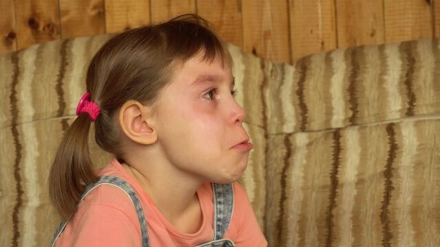 Little child, upset and crying girl of 6-7 years old against the background of a wooden wall in a summer house in the village. A girl of Russian or European appearance with funny pigtails.