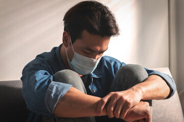 An upset health problem Asian patient man wearing a face mask at home or hospital alone sad on a sofa waiting for a doctor. Hospital and Health care during Coronavirus or Covid-19 quarantine concept