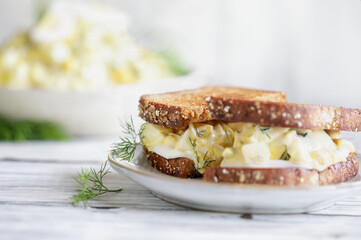 Abstract of an egg salad sandwich with fresh dill over a rustic wood table. Extreme selective focus with blurred foreground and background with bowl of spread in back.