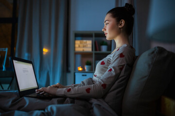 technology, bedtime and people concept - teenage girl with laptop computer sitting in bed at home...