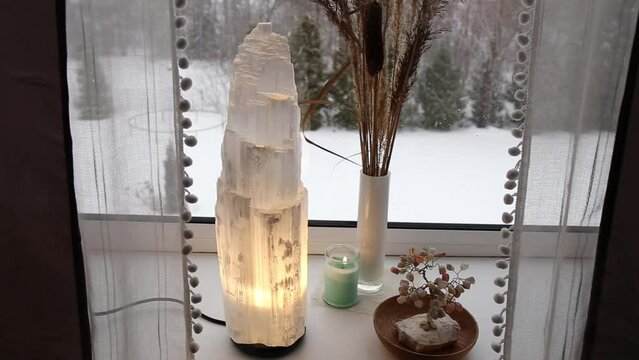 Switch on rough big selenite crystal tower pole lamp illuminated on home window sill, spiritual home decor accent. Winter forest on background.