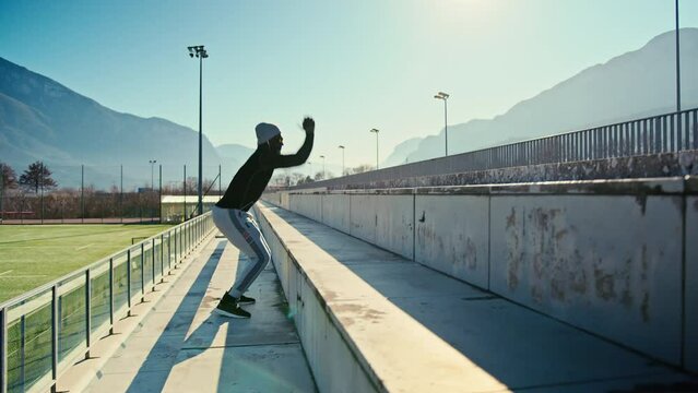 Athletic black man is jumping up the tribunes of an outdoor stadium on a sunny day. He is wearing unbranded sports clothes and mirrored sunglasses. Beautiful mountain landscape on a background.