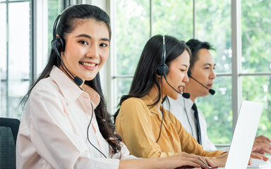 Focus Asian beautiful attractive female call center cheerful smiling with headsets, sitting, looking camera, monitoring with colleagues in office or agency at computer, giving service to customers.