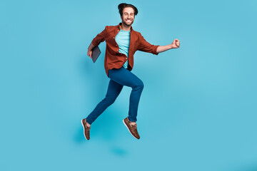 Fototapeta na wymiar Full length photo of cool brunet millennial guy run with laptop wear eyewear shirt trousers sneakers isolated on blue background