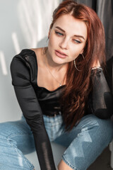 Beautiful young girl in a fashionable denim outfit with a black T-shirt and blue jeans is sitting near the window with sunlight indoors