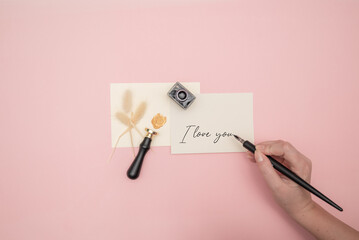 Top view of female hand writing I Love You note on pink background.  Note with phrase I love you, top view. Space for text