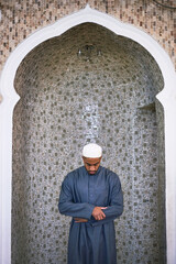 A young Muslim man stands with his head bowed in prayer in a mosque with a mosaic tile background