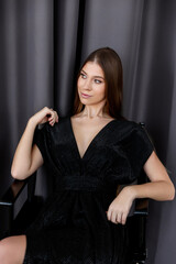 A gorgeous and sexy brunette Caucasian woman in a black dress poses on a chair. Long and healthy hair, bright and glowing skin. Beautiful cleavage of the dress.
