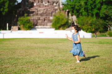 Cute Asian kid girl is running on grass in warm sunshine. Children wear face mask to prevent PM2.5...