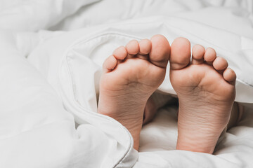 Childs legs are covered with white bed linen. The warmth of home comfort.