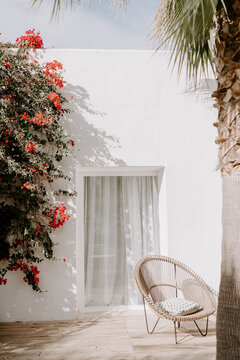 A serene Ibiza-style patio with a white stucco wall, sheer curtains, a woven rattan chair, and vibrant bougainvillea, creating a peaceful Mediterranean retreat bathed in natural sunlight