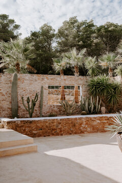 Charming Mediterranean patio in Ibiza featuring rustic stone walls, modern seating, and a tall cactus. A serene outdoor escape blending nature and architecture in a stylish bohemian setting