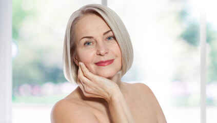 Happy beautiful middle-aged blonde woman shows off her perfectly well-groomed face and sitting relaxed near the window. Realistic images.
