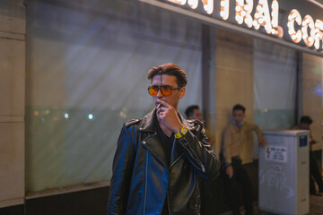 Portrait of young handsome brunet caucasian man in black leather jacket and yellow sunglasses. Standing on the street at night and smoking.