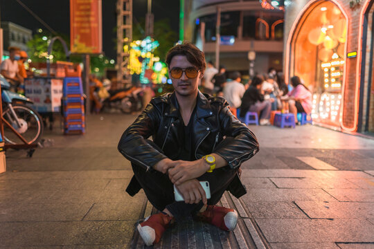 Portrait of young stylish brunet caucasian man in black leather jacket and yellow sunglasses Sitting on the street at night. High quality photo