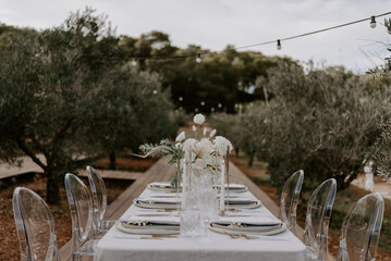 Elegant outdoor destination wedding table setup with white linens, floral centerpieces, transparent chairs, and string lights, creating a romantic and intimate atmosphere for a special celebration