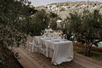 Elegant outdoor destination wedding table setup with white linens, floral centerpieces, transparent chairs, and string lights, creating a romantic and intimate atmosphere for a special celebration