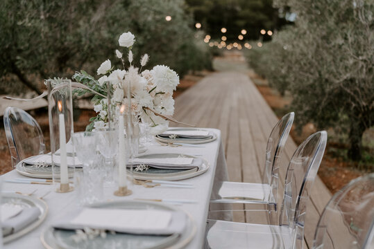 Elegant outdoor destination wedding table setup with white linens, floral centerpieces, transparent chairs, and string lights, creating a romantic and intimate atmosphere for a special celebration