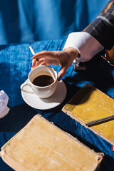 Fototapeta na wymiar partial view of woman with cigarette near cup of coffee and vintage books on blue velour tablecloth