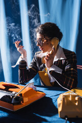 retro style journalist with cigarette talking on telephone near typewriter on blue background