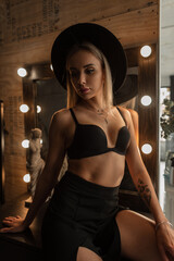 Fototapeta na wymiar Hot sexy young blond woman model with a cute face and slender body in a vintage elegant hat with a black bra and a dress sits near a mirror with light bulbs indoors