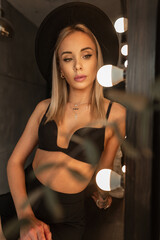 Fototapeta na wymiar Fashion portrait of a beautiful sexy woman with an elegant hat in a black bra sits near a mirror with light bulbs