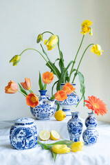 A Lush natural light stillife with old Dutch Delft blue lidded vase with yellow and orange tulips a gerbera and lemons on white  linen against a white background.