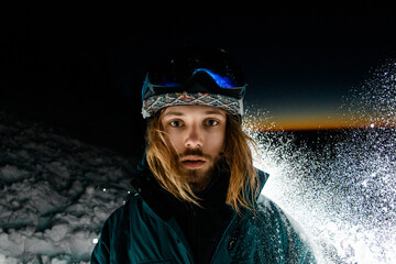 portrait of blonde man in ski helmet on background of dark evening sky and backlit splashing snow.