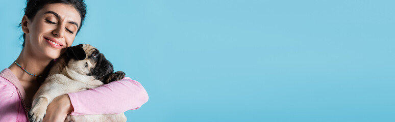 happy brunette woman with closed eyes embracing pug dog isolated on blue, banner