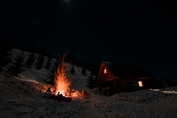 great view of wooden house and high bonfire on snow-covered mountainside at night
