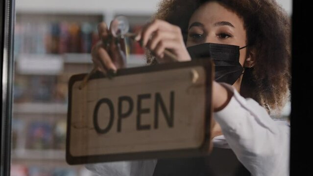 Young african american woman waiter in medical mask getting ready for start working day cafe owner turning sign that says OPEN welcomes new customers in modern coffee shop girl bartender opens bar