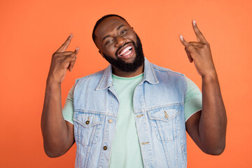 Photo of cool funny afro guy wear denim vest showing hard rock sign isolated orange color background