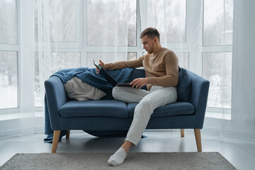 A young man works at home on the couch with his laptop. A man with a mobile phone and a laptop is...