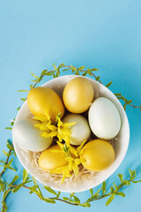 Yellow and white eggs in plate decorated with willow branches and flowers on a pastel blue background, Easter decor. Flat lay, top view. Copy space for text.	