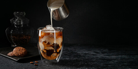 Iced coffee with pouring cream on dark stone background. Copy space