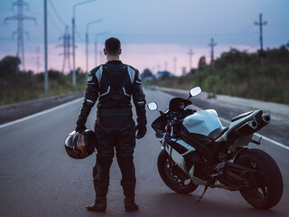 A biker stands next to a motorcycle on an asphalt road and looks into the distance. Summer sunset © pavelkant