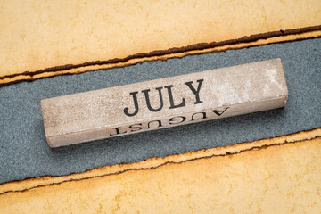 July text on grunge wooden block against handmade rag paper in gray and yellow tones, calendar...