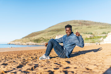 Fototapeta na wymiar young african american man sitting on the beach smiling raising thumb up on a sunny day. approval concept