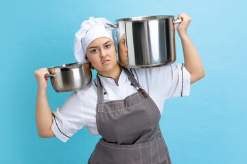 Portrait of young annoyed woman, lady-cook, chef in white uniform with saucepans isolated on blue...