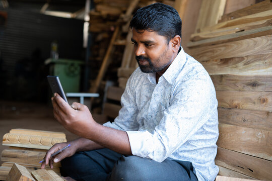 smiling carpenter busy using mobile phone at carpentry shop - concept of technology, social media, surfing internet and checking deisgns