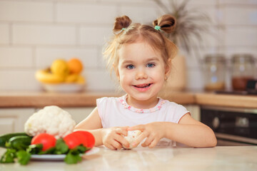 Obraz na płótnie Canvas A happy girl 2-3 years old in the kitchen at home or in kindergarten eats delicious and healthy vegetables for lunch.