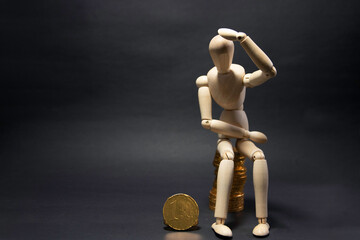 Model of a wooden figure sitting on a pile of coins being confused, holding a coin in his arms on a black background. Business concept