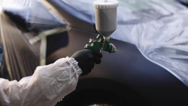 Close-up of manual spraying machine with paint for car is painting metal detail. Worker is holding and directing equipment in car service center. Modern car painting