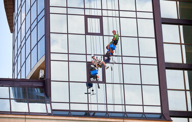 Industrial mountaineering workers washing glass windows of high-rise building, hanging on safety climbing ropes. Two men window cleaners working together outside skyscraper.
