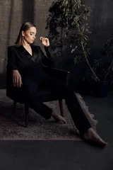 Beautiful brunette sitting in a black suit. looking down. Back gray background and flowerpot © monchak