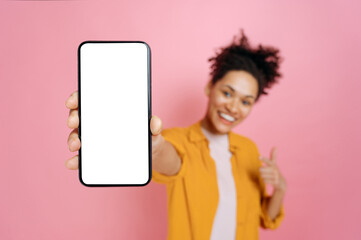 Smartphone mock-up concept. Defocused african american girl, showing cellphone with blank white screen for presentation, stand over isolated pink background, smiles joyfully. Copy-space, advertisement