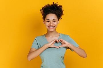 Lovely candid pleasant african american young woman, in basic t-shirt, makes heart gesture with hands, demonstrates love sign, stands on isolated orange background, looking at camera, smiling friendly